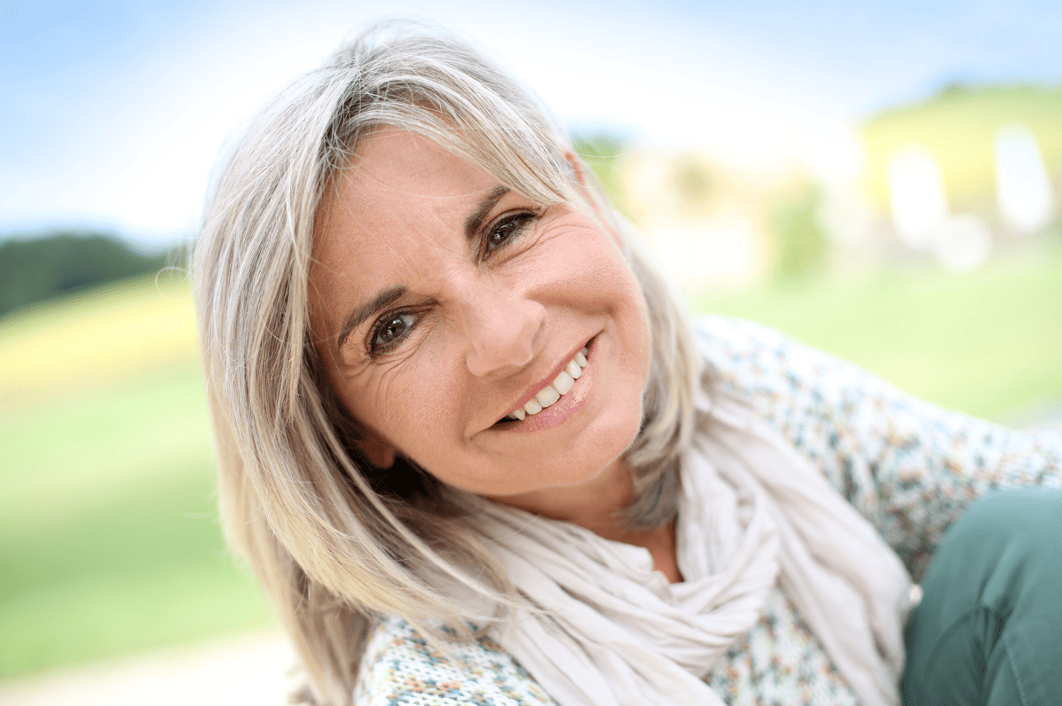 Eine Frau mit grauen mittellangen Haaren sitzt lächelnd vor einem hellgrünen Hintergrund.