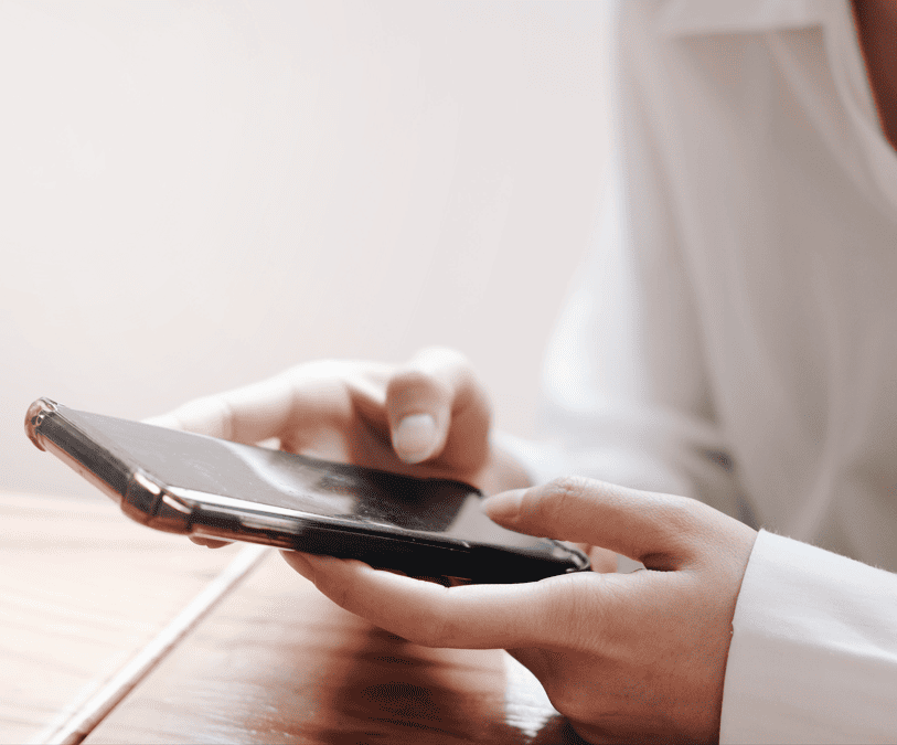 Eine Frau mit weisser Bluse hat ein Handy in beiden Händen und tippt mit den beiden Daumen darauf.